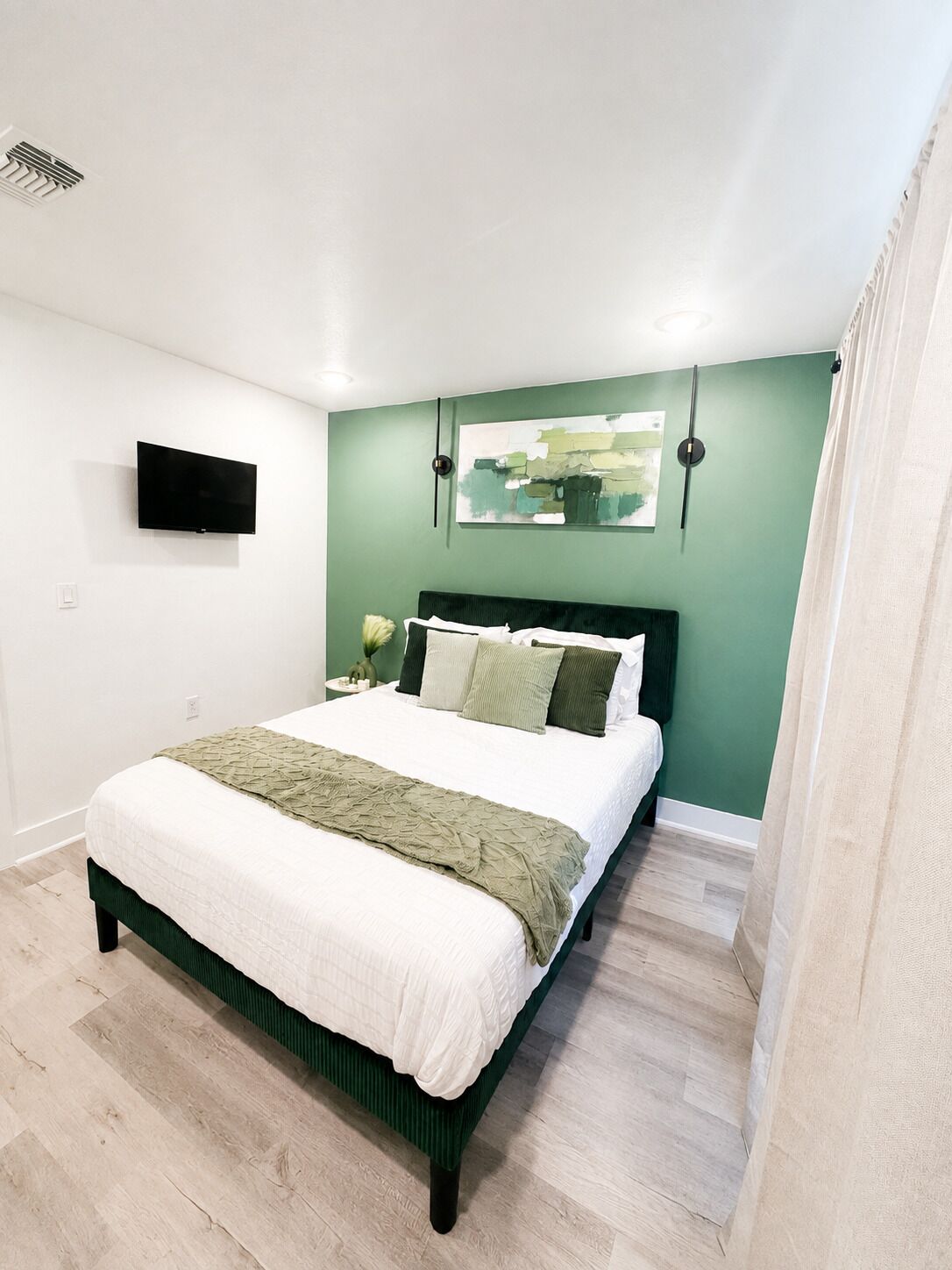 A bedroom with a bed, a flat-screen TV mounted on the wall, and a painting above the bed.