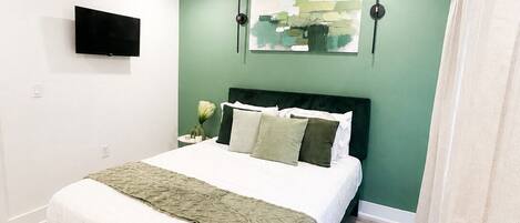 A bedroom with a bed, a flat-screen TV mounted on the wall, and a painting above the bed.