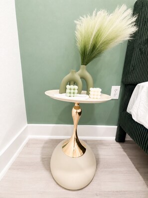 A small round table with a gold base, a vase with green plants, and two decorative items on a light-colored surface against a green wall. - The Sage Stay Modern Retreat In Downtown Dallas (Dallas)