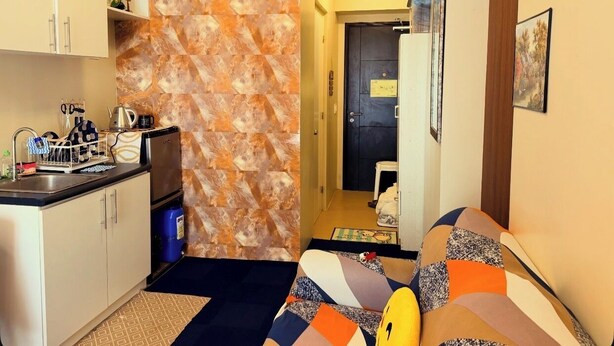 Een ruimtelijk ingerichte leefruimte met een kitchenette, een behang met patroon, een bank en een deur naar een andere kamer.