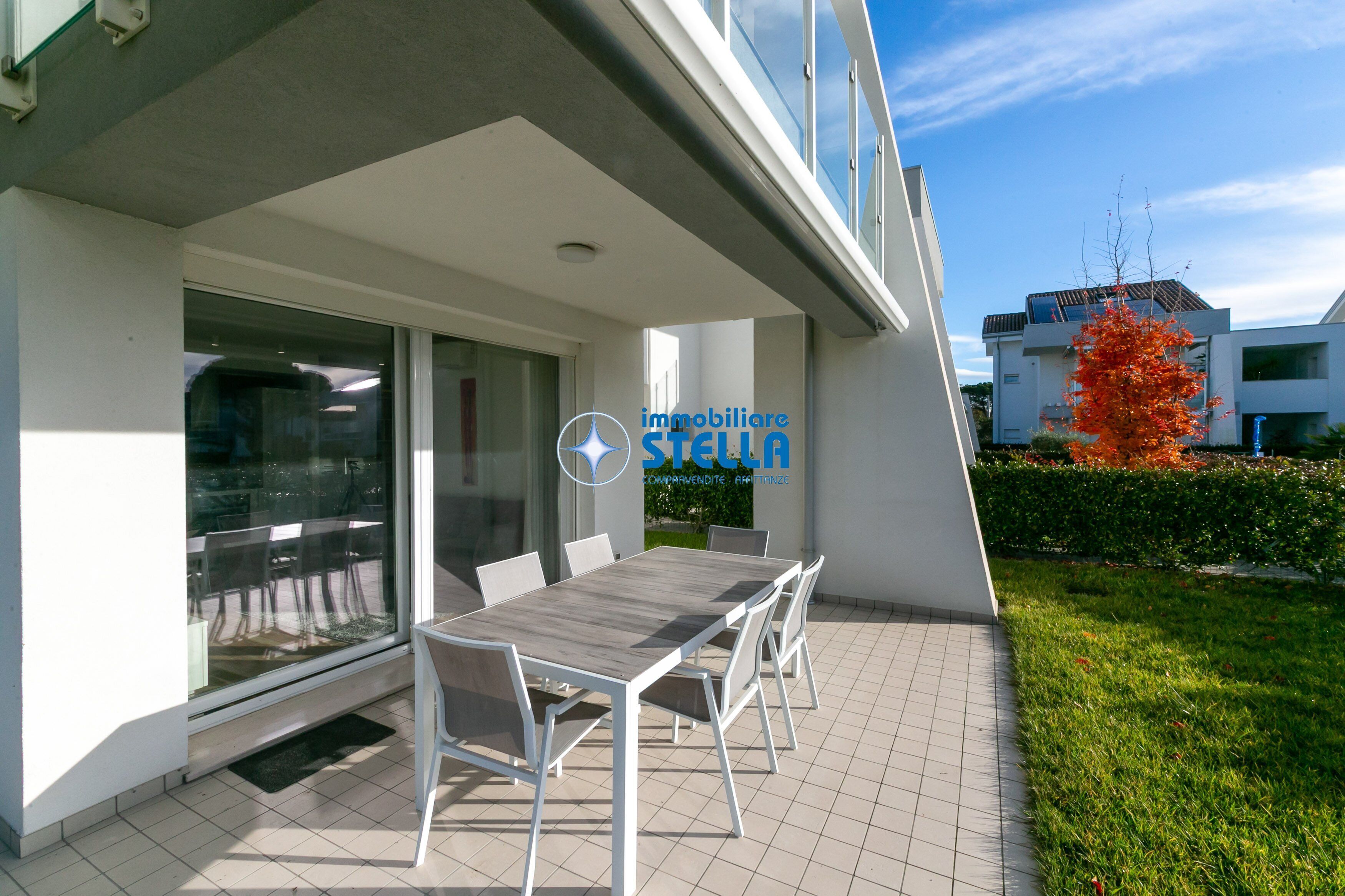 Een modern buitenpatio met een eettafel en stoelen, een glazen toegang en het logo van 'immobiliare STELLA'.
