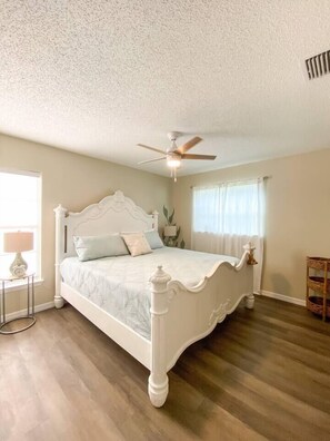 Une chambre à coucher avec un lit blanc, un ventilateur de plafond, une fenêtre avec des rideaux et une petite table avec une lampe.