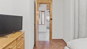 Un dormitorio con una cama, un mueble de televisión de madera con un televisor de pantalla plana, una puerta de madera que da al baño y piso de baldosas.