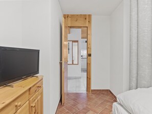 Ein Schlafzimmer mit einem Bett, einer Holz-TV-Kommode mit Flachbildfernseher, einer Holztür zu einem Badezimmer und einem Fliesenboden.