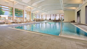Una piscina interior con suelo de baldosas, sillas de descanso y ventanas amplias.
