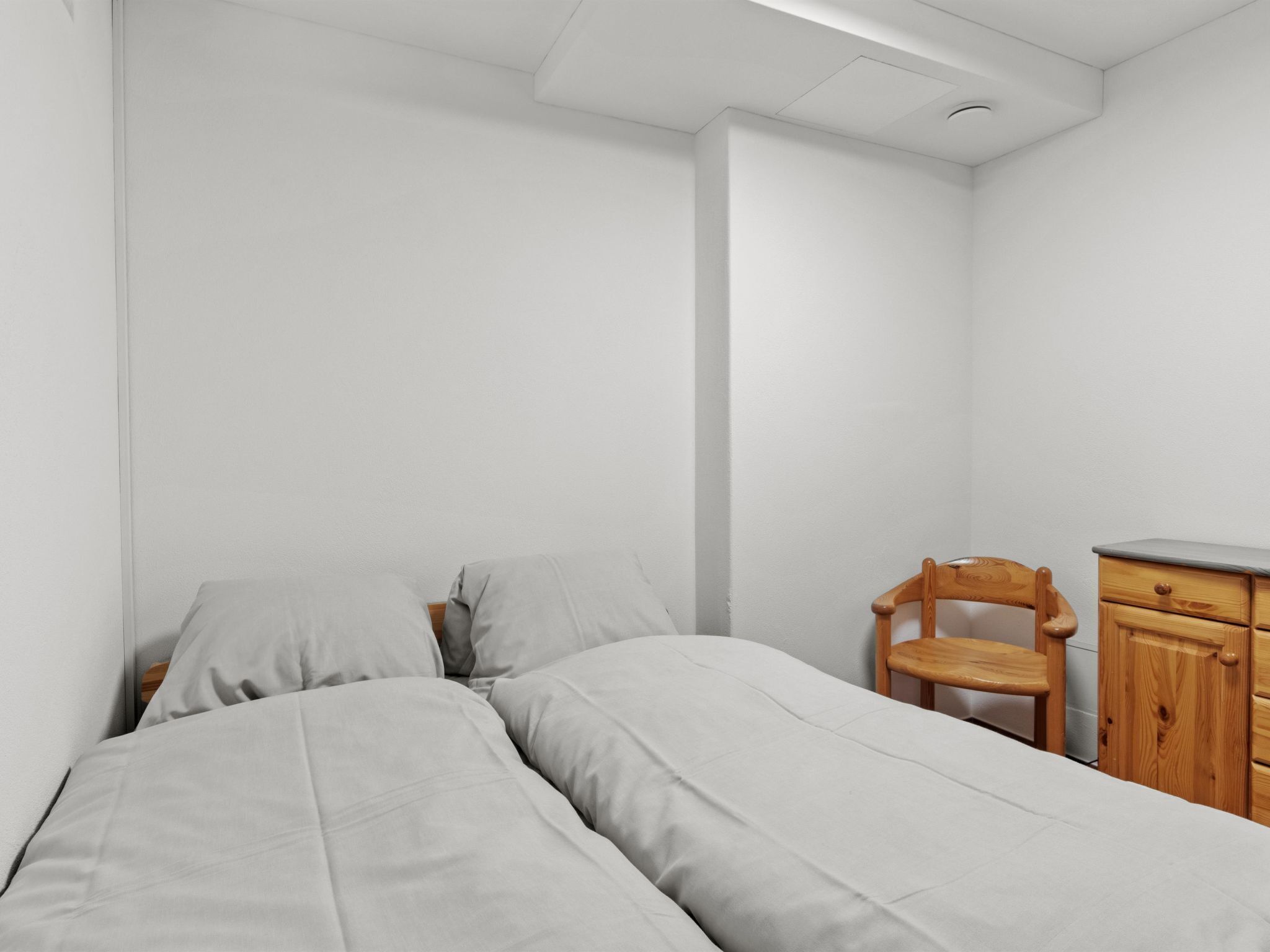 A simple, unadorned room with two beds, a wooden chair, and a small wooden cabinet.