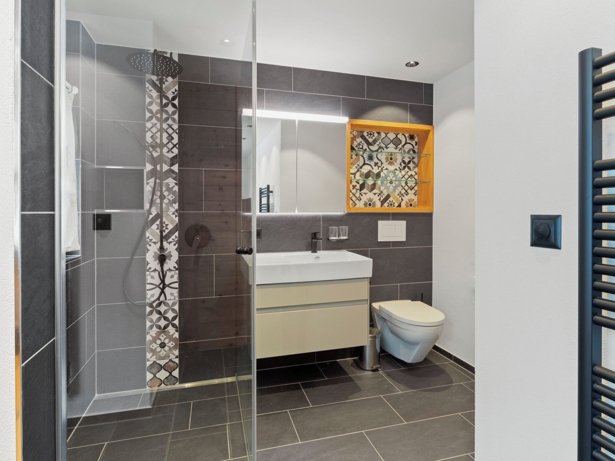 Een badkamer met een douchecabine van glas, een witte wastafel, een toilet en een spiegel met een decoratieve tegelachtergrond.