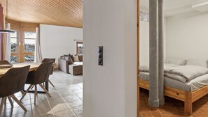 Un interior moderno con techo de madera, un comedor con sillas y mesa, y un dormitorio con cama y dos columnas.