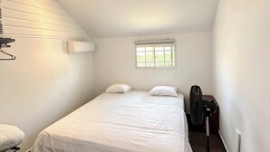 Ein kleines, helles Schlafzimmer mit einem Einzelbett, einem Fenster und einem Standventilator.
