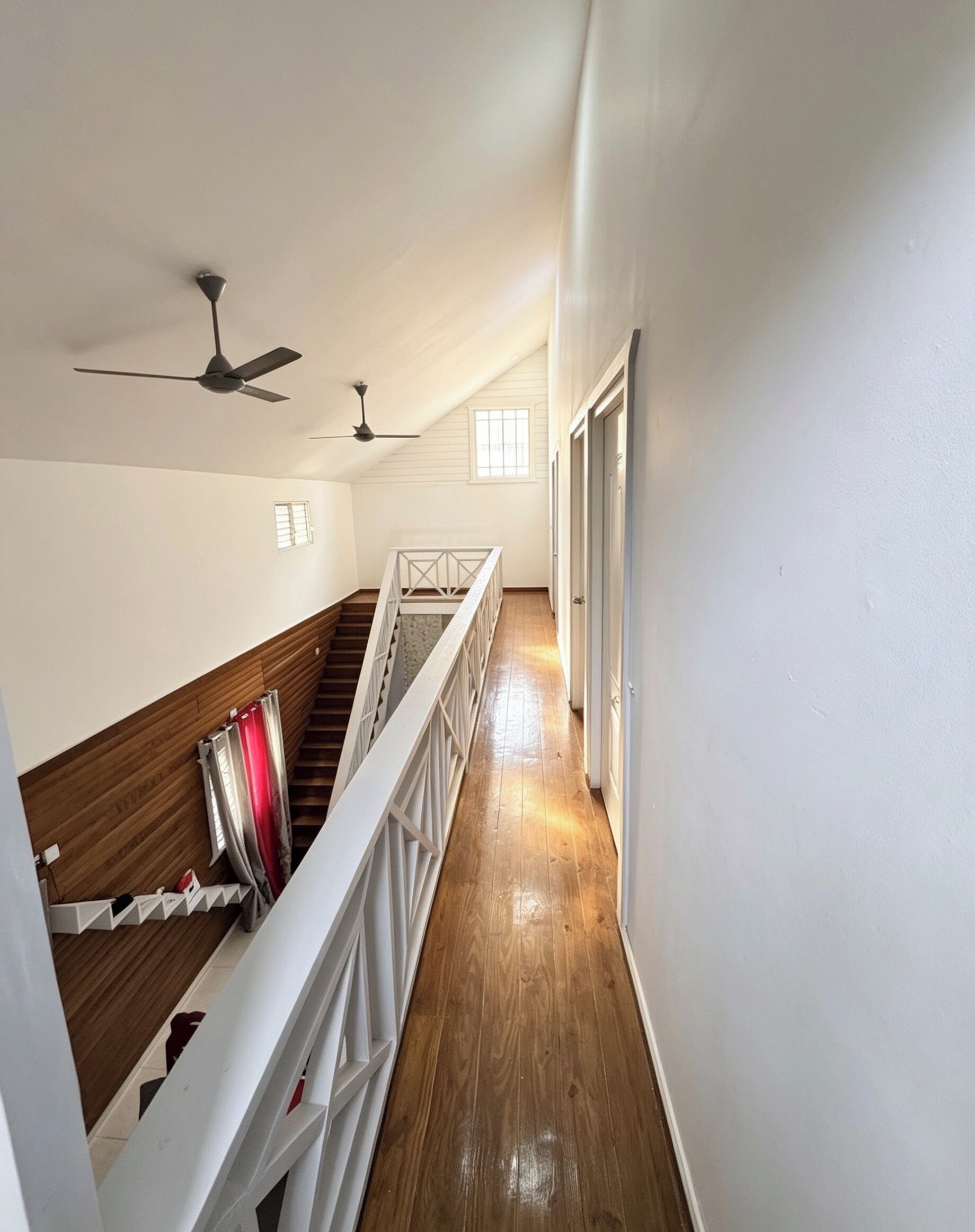 A hallway with wooden flooring, white walls, and a staircase with a railing. There are two ceiling fans and a window allowing natural light.