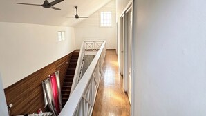Ein Flur mit Holzboden, weißen Wänden und einer Treppe mit Geländer. Zwei Deckenventilatoren und ein Fenster sorgen für natürliches Licht.