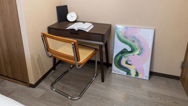 Een compact werkplekje met een bureau, stoel, klok, open boek en een kleurrijk abstract schilderij.