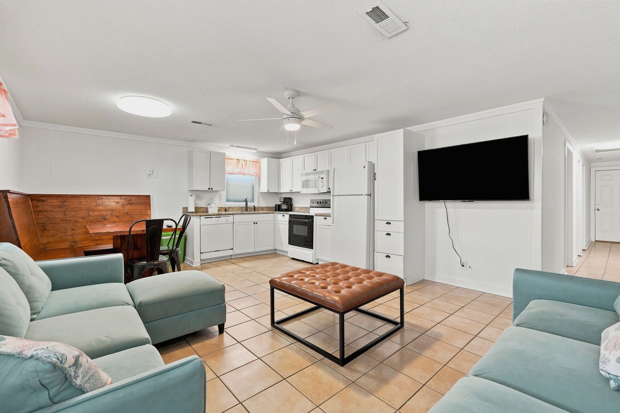 A modern living room with a brown leather ottoman, teal sofas, and a flat-screen TV mounted on the wall.