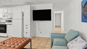 A compact living space with a kitchenette, a flat-screen TV mounted on the wall, a brown leather bench, and a teal sofa with floral cushions. - Cherry Grove Coastal Escape · 2BR · Steps to Beach (North Myrtle Beach)