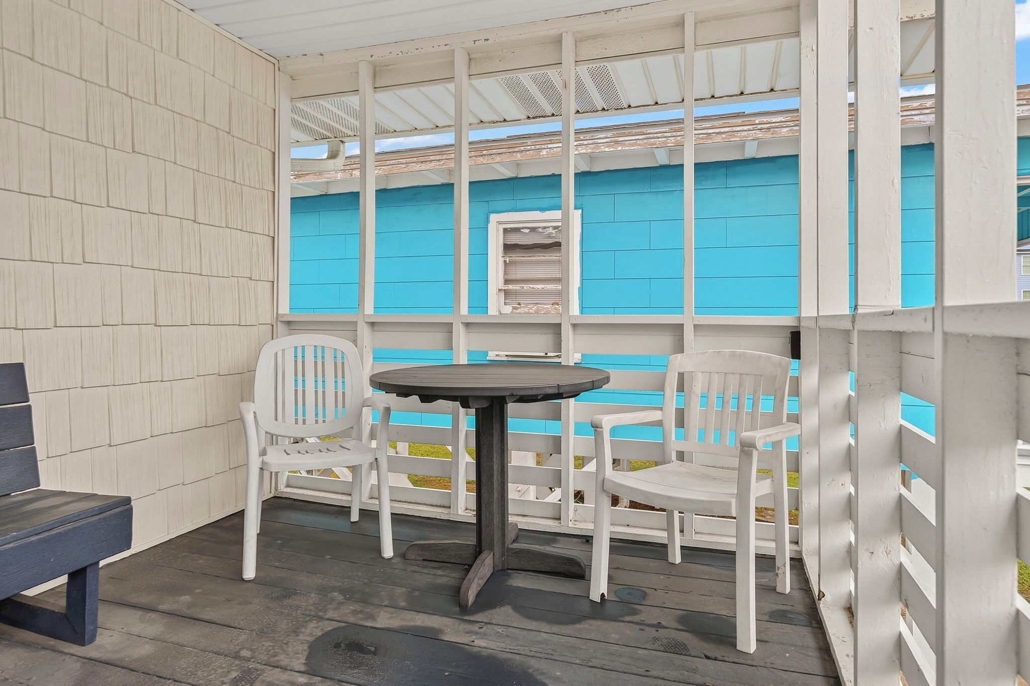 A covered outdoor seating area with a table and chairs, a bench, and a view of a blue building.