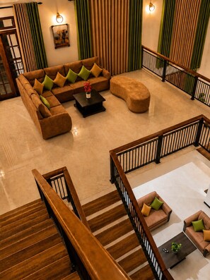 Un vaste espace de vie comprenant un canapé d’angle, un pouf et une table basse, un escalier avec des rampes en bois, et une vue sur l’extérieur grâce à de grandes fenêtres.