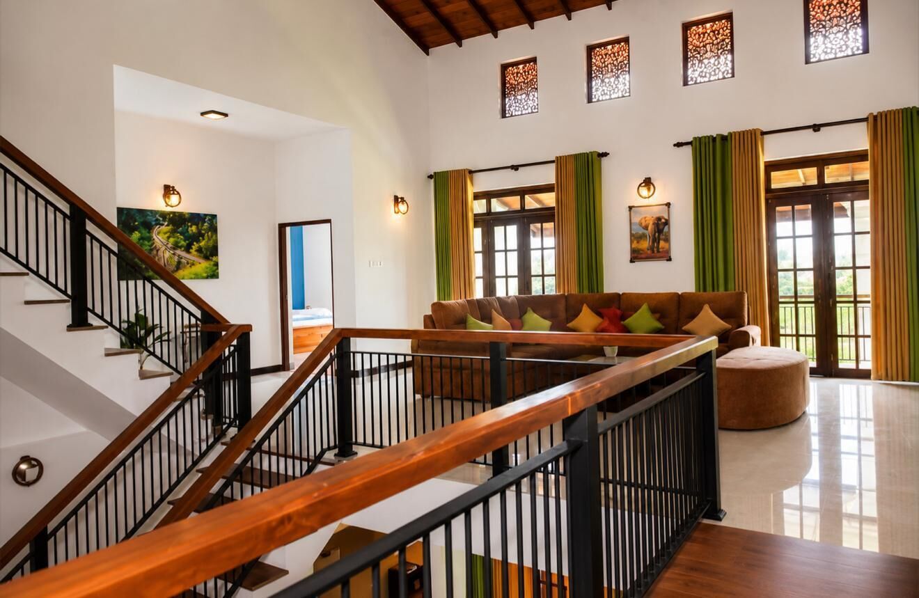 A spacious living area with a wooden staircase, a sofa with colorful cushions, and a large window with green curtains.