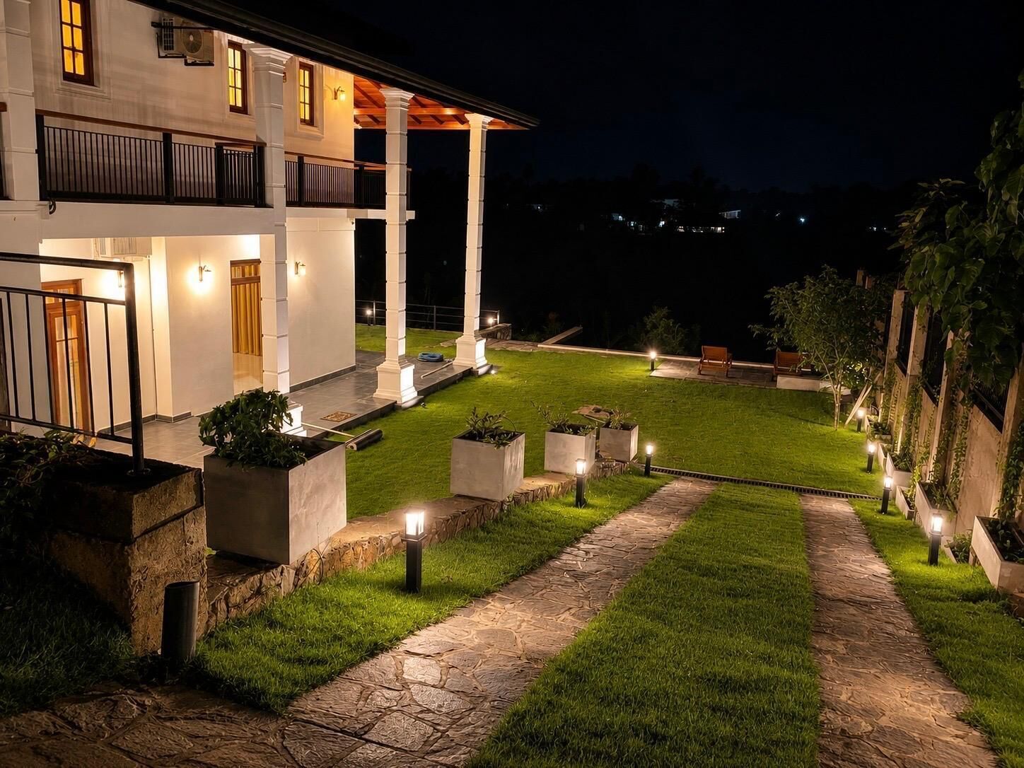 A well-lit outdoor area with a paved walkway, planters, and a building with multiple windows and a balcony.