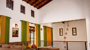 A spacious living room with a brown sofa, a curved ottoman, and a coffee table. There are green and yellow curtains, a framed picture on the wall, and a balcony with a railing. - Luxury 5BR Family Villa Near Airport, Pool, Rooftop, Sleeps 14+ (Pansilgoda)