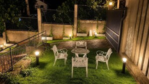 An outdoor patio area with a stone table and four chairs, surrounded by a well-lit garden with grass and plants. - Luxury 5BR Family Villa Near Airport, Pool, Rooftop, Sleeps 14+ (Pansilgoda)