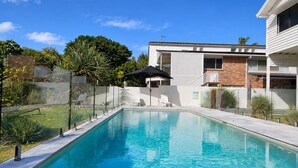 Una piscina rectangular con superficie azul cristalina, rodeada por una valla blanca, y un edificio con balcón al fondo.