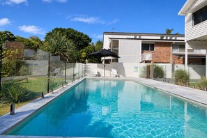 A rectangular swimming pool with a clear blue surface, surrounded by a white fence, and a building with a balcony in the background. - Sea Breezes and Sand Between Your Toes (Sunshine Coast)