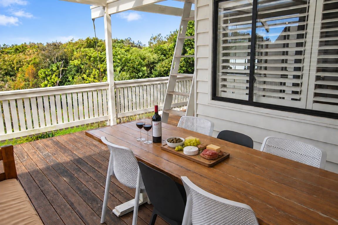 Une terrasse extérieure en bois, avec une table dressée pour un repas, des chaises blanches et une vue sur la verdure.