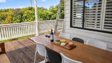 Une terrasse extérieure en bois, avec une table dressée pour un repas, des chaises blanches et une vue sur la verdure.