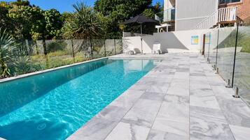 A swimming pool with clear blue water, surrounded by a tiled deck and a glass fence.