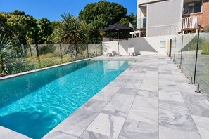 A swimming pool with clear blue water, surrounded by a tiled deck and a glass fence.