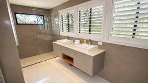Un baño moderno con cabina de ducha de vidrio, tocador doble y ventana con persianas.