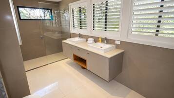 Une salle de bain moderne avec une douche à l’italienne en verre, un meuble-lavabo double et une fenêtre avec des stores.