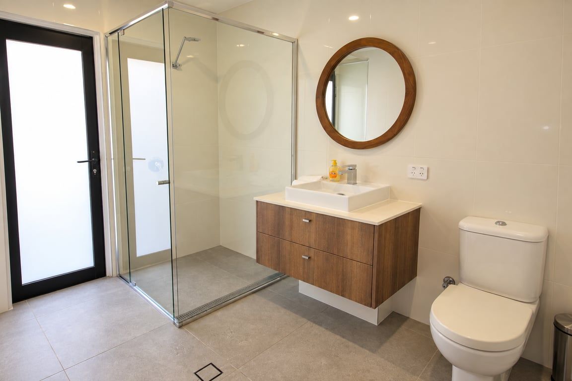 Ein modernes Badezimmer mit Duschkabine aus Glas, Waschtisch aus Holz, rundem Spiegel und weißer Toilette.