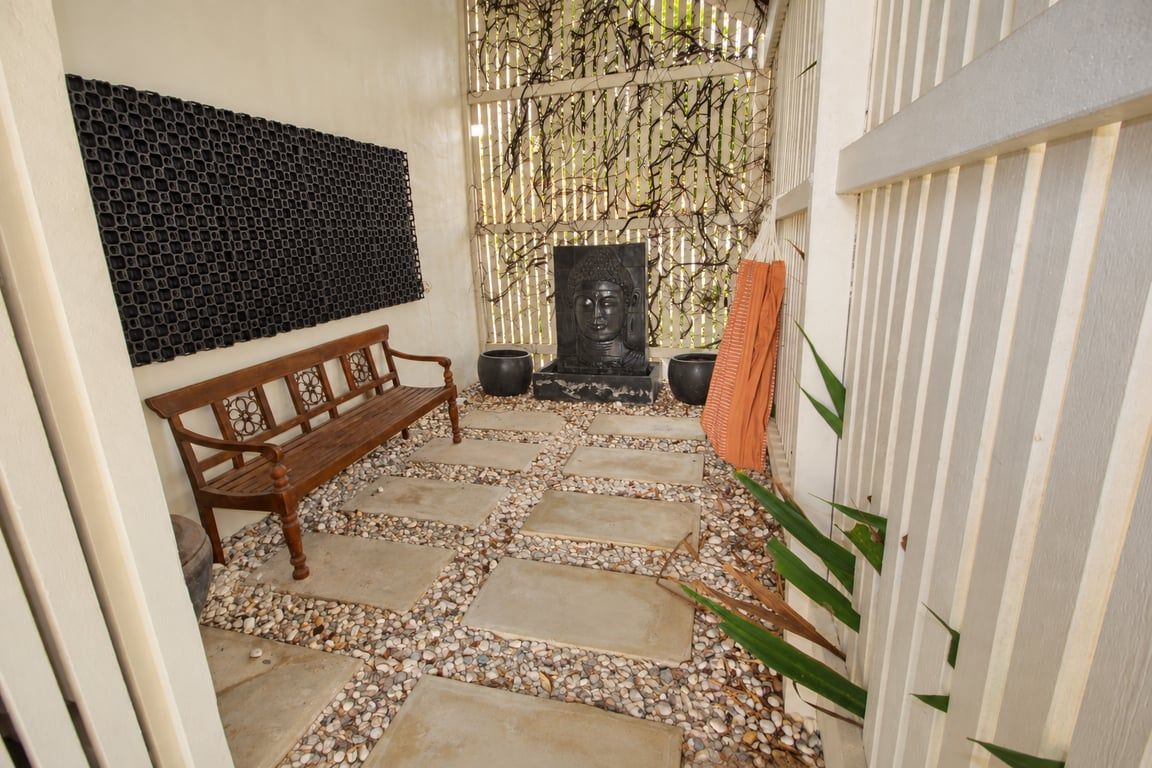 Un sentier dallé de pierres mène à un banc en bois et à une statue de Bouddha, avec un mur décoratif et une clôture blanche.