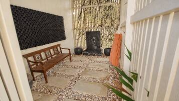 Un sentier dallé de pierres mène à un banc en bois et à une statue de Bouddha, avec un mur décoratif et une clôture blanche.