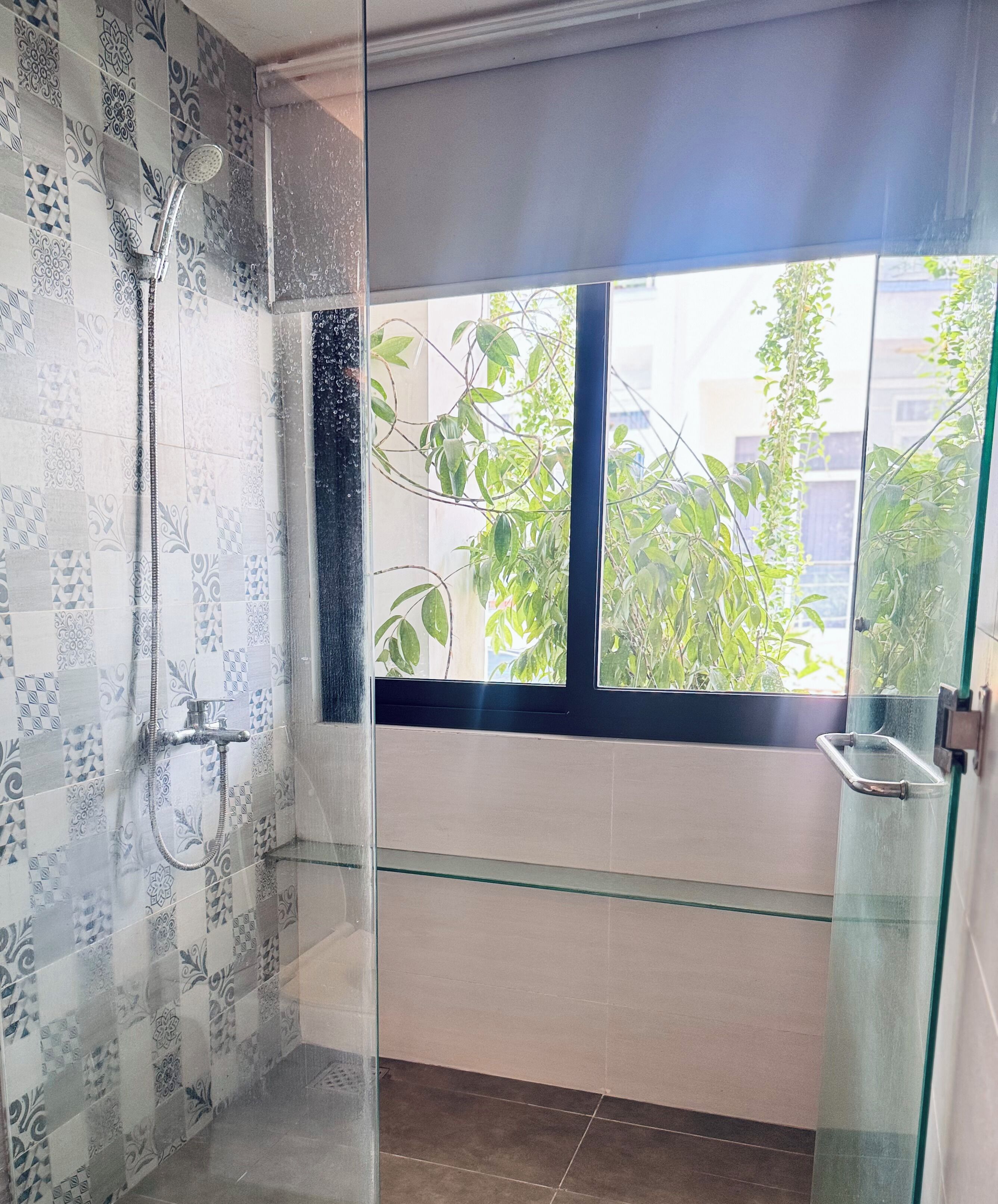 Un área de ducha con paredes de vidrio, baldosas con diseño, una puerta de vidrio y una ventana con vista a follaje.