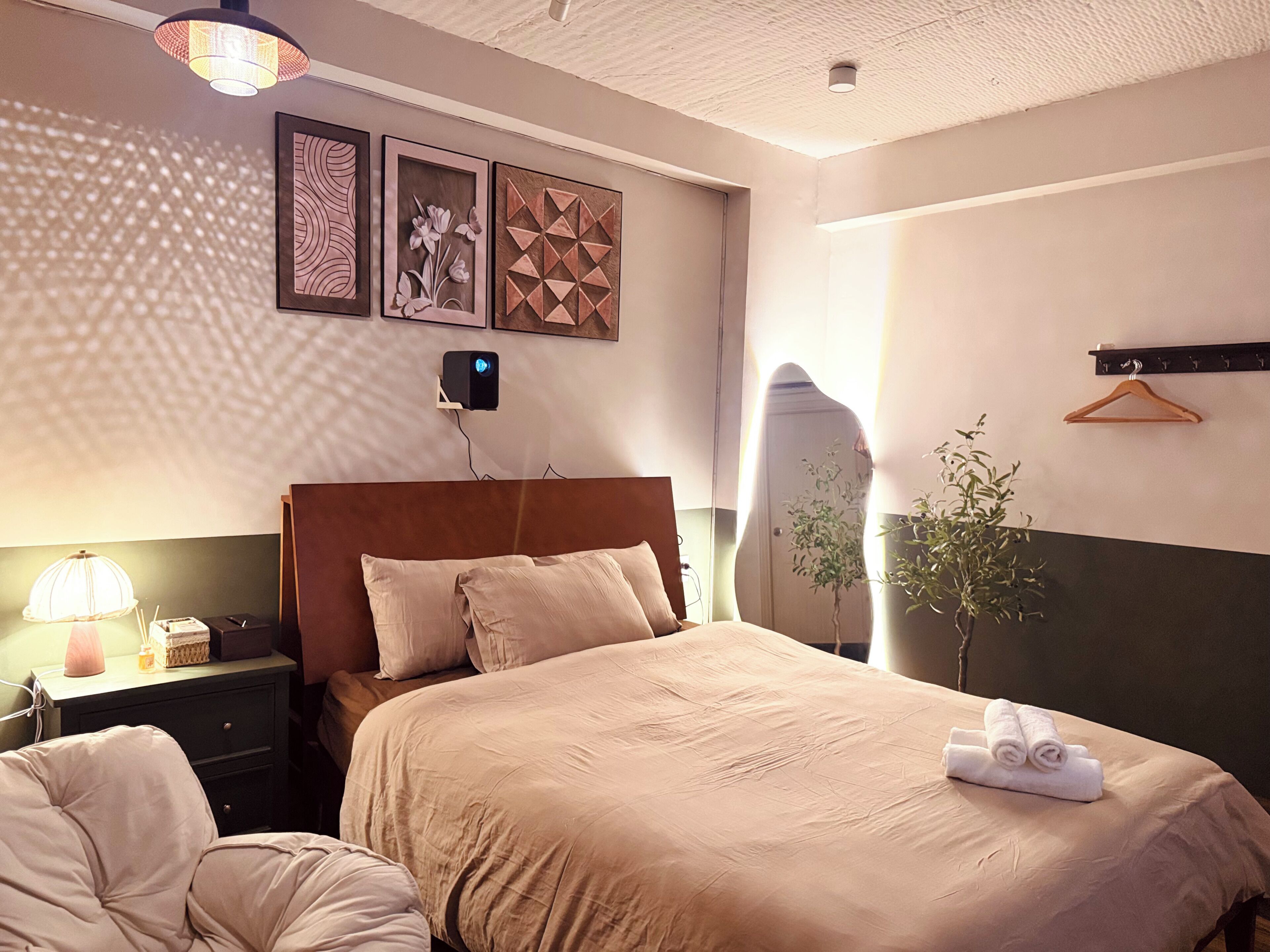 Hotellihuone, jossa on sänky, yöpöytä, lamppu, seinätaide ja roikkuva kasvi.