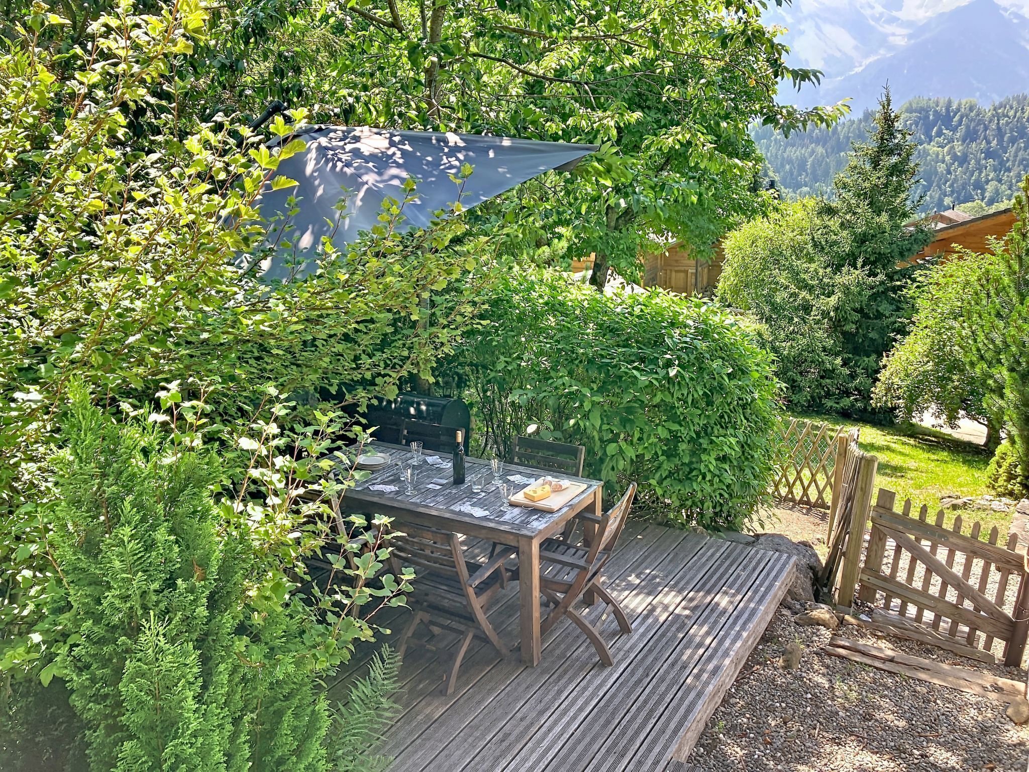Une terrasse en bois avec une table dressée pour le repas, entourée d’une végétation luxuriante et offrant une vue sur les montagnes en arrière-plan.