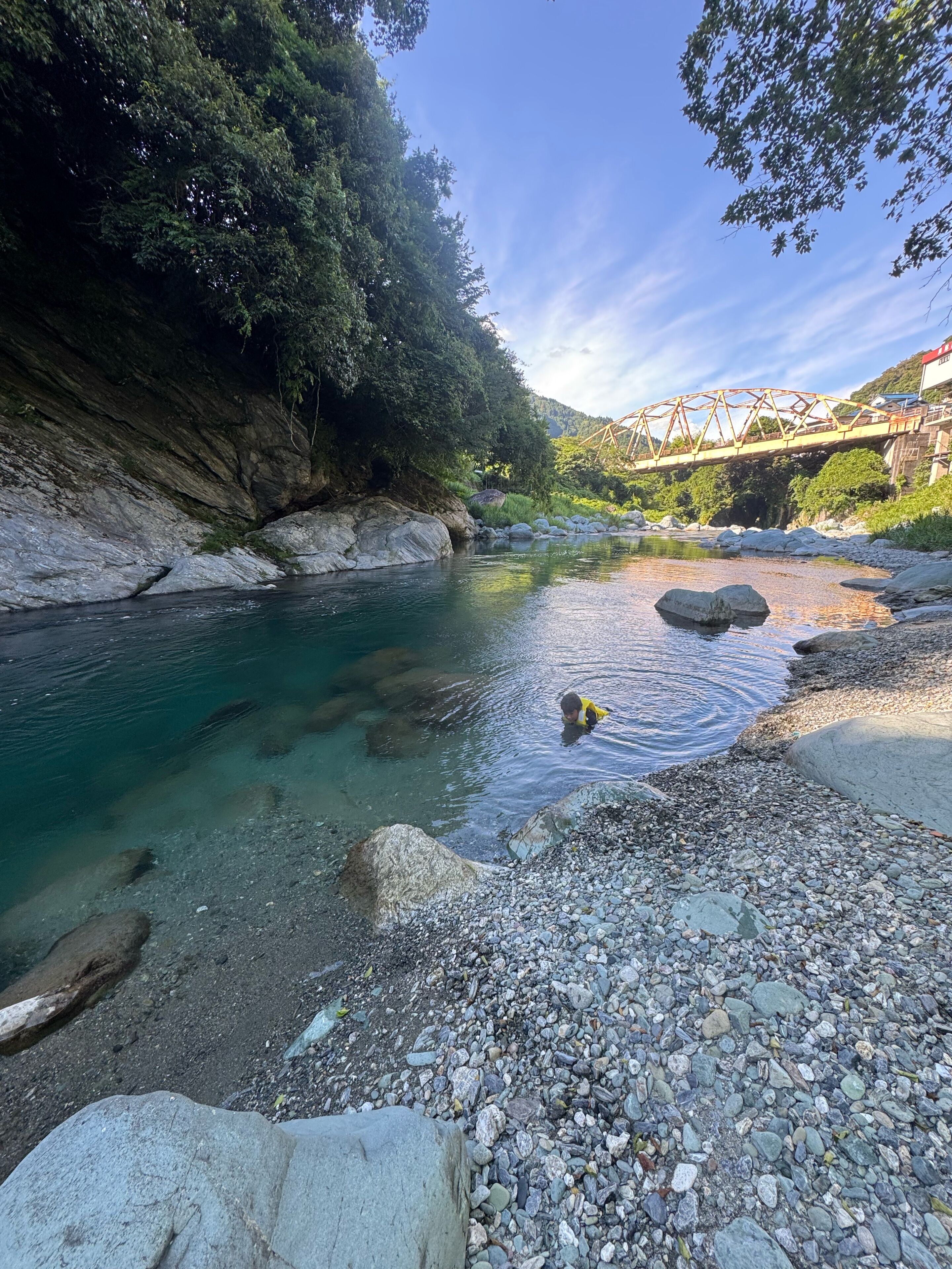 A river with clear water, surrounded by rocks and greenery, with a bridge in the background.