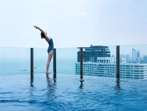 Una persona con traje de baño azul realizando una postura de yoga en una piscina infinita con fondo de cristal y un horizonte urbano al fondo.