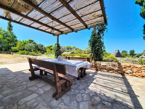 Ein Außenbereich mit Tisch und Bänken unter einer Holzpergola, umgeben von Steinmauern und Grünpflanzen.