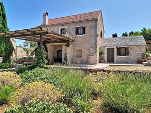 Ein Steinhhaus mit Ziegeldach, einer Holzpergola und einem Garten mit verschiedenen Pflanzen.