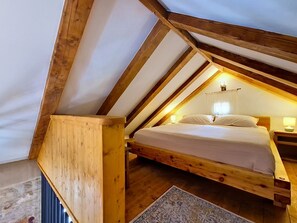 Ein Schlafzimmer mit schräg versiegelter Decke, Holzbalken, einem Bett, einem Nachttisch und einer Wandleuchte.