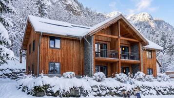 A two-story wooden chalet with a balcony, set against a snowy mountain backdrop.