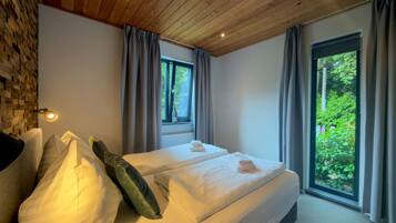 A modern bedroom with a wooden ceiling, a large bed with white linens, and a view of greenery through the windows.