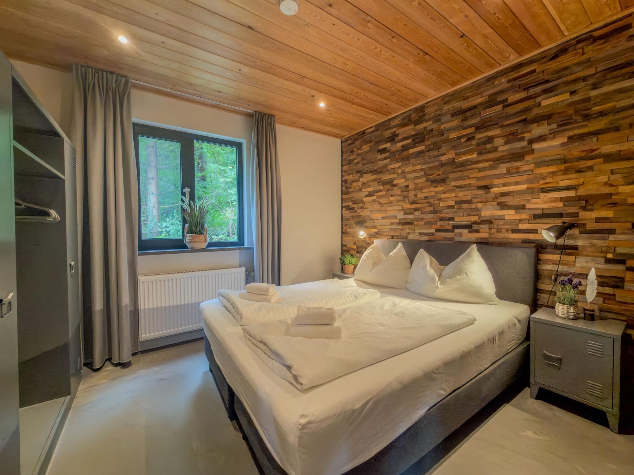 A bedroom with a wooden ceiling, a large bed with white bedding, a nightstand with a lamp, a window with curtains, and a radiator.