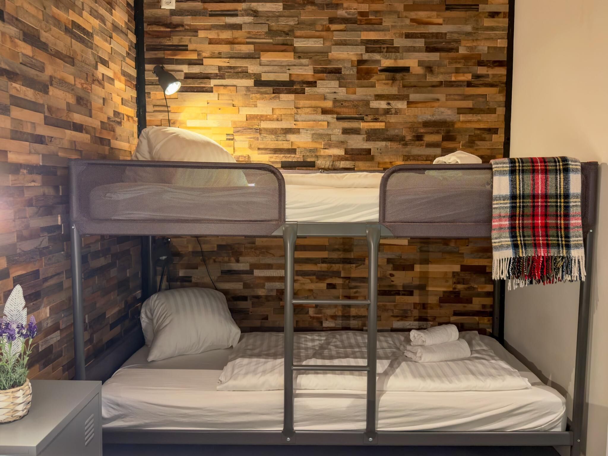 A bunk bed room with a wooden wall, a desk with a lamp, and a plaid blanket on the right.