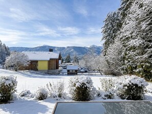 A snow-covered landscape with a wooden cabin, a small pool, and snow-laden trees.