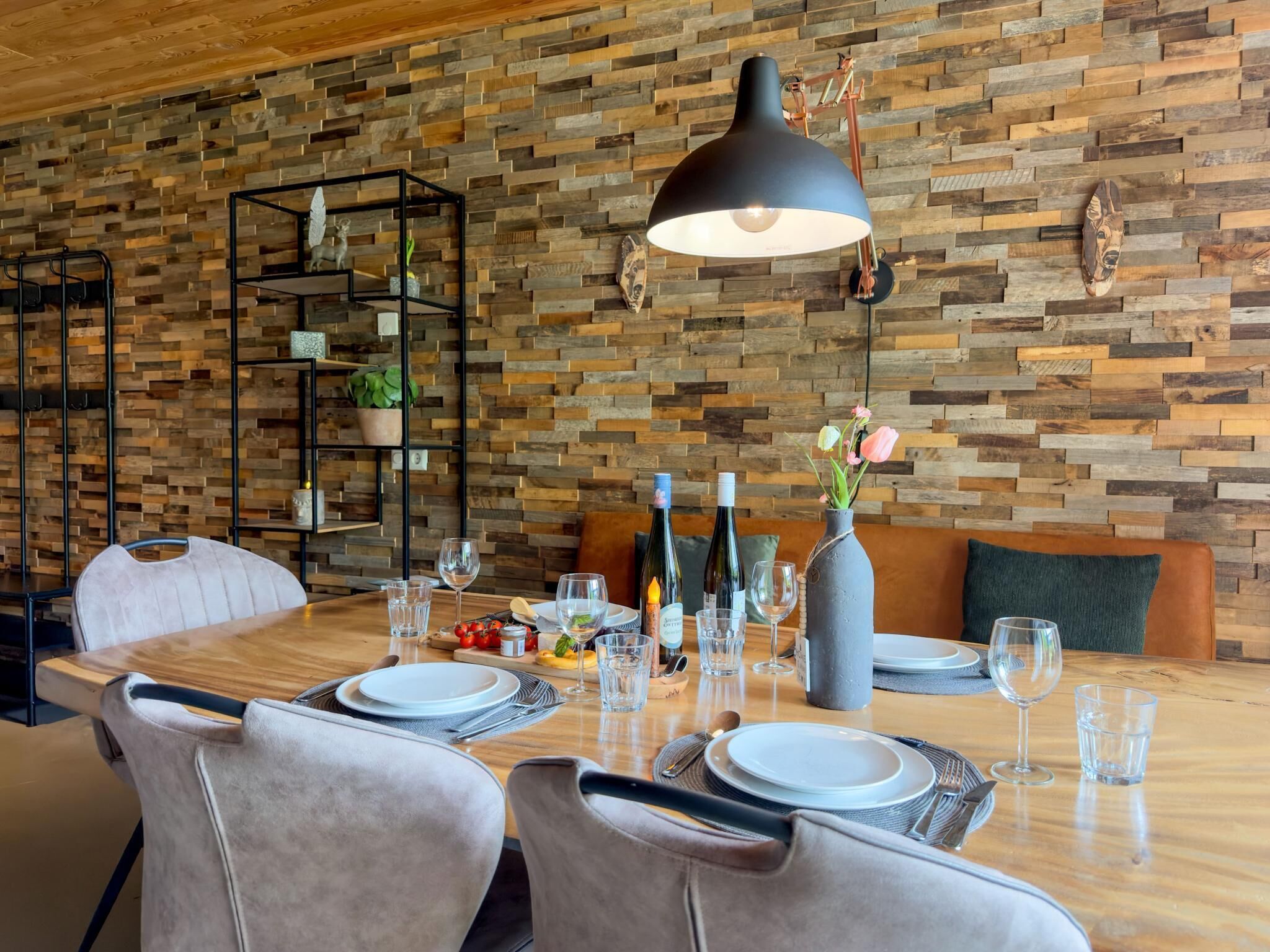 A dining area with a wooden table set for two, surrounded by chairs. A black pendant light hangs above the table, and a wooden wall with a herringbone pattern serves as the backdrop.