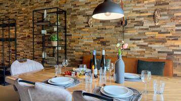 A dining area with a wooden table set for two, surrounded by chairs. A black pendant light hangs above the table, and a wooden wall with a herringbone pattern serves as the backdrop.